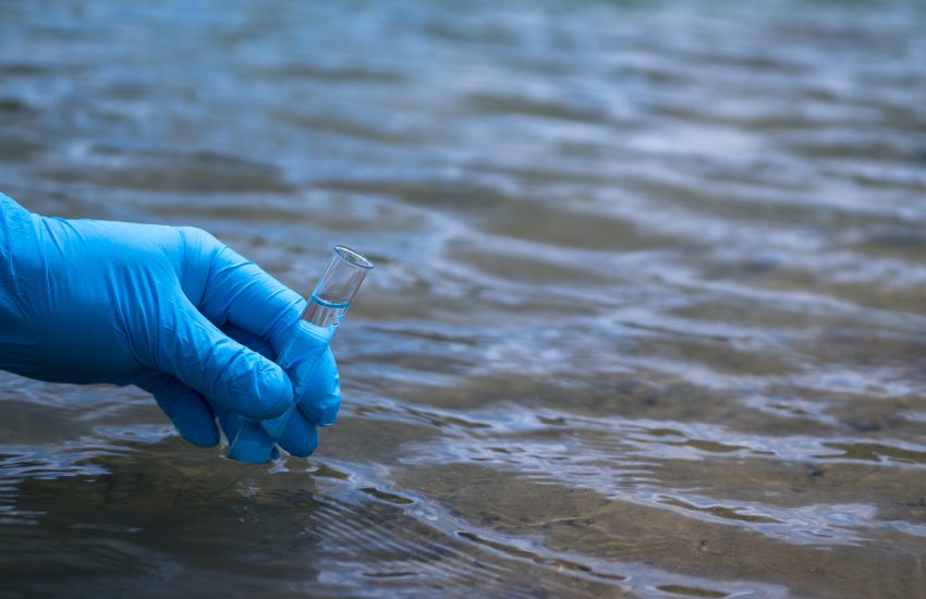 A man's hand in a blue glove takes a close-up of water into a test tube to measure water pollution. Background